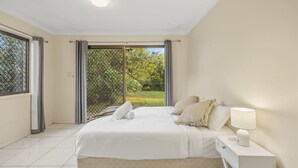 A bedroom with a bed, a bedside table with a lamp, and a view of greenery through a window.