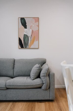 Een grijze bank met kussens, een lijst met een abstract schilderij en een witte stoel in een kamer met houten vloeren.