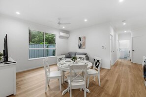 Een moderne keuken met witte kasten, een eethoek met een ronde tafel en stoelen, en een grijze bank op de achtergrond.