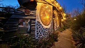 Une cabane rustique dotée d’une grande fenêtre circulaire, de murs en bois et d’un sentier menant à l’entrée.
