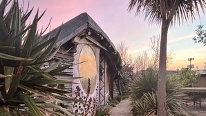 Un bâtiment rustique doté d’une grande fenêtre circulaire, entouré d’une végétation tropicale et bordé d’un chemin pavé.