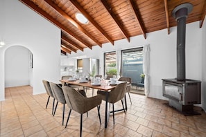 A dining area with a wooden table and chairs, a kitchen in the background, and a wood-burning stove.