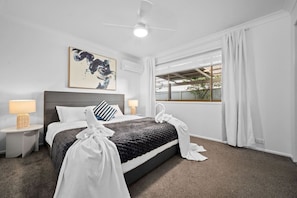 A neatly arranged hotel room with a bed, two bedside tables, a ceiling fan, a window with curtains, and a wall-mounted air conditioner.