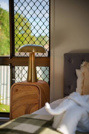 A bed with a headboard, a bedside table with a lamp, and a window with a metal screen. - Cozy Self-Contained |Hospital, Train & Shop Nearby (Loganlea)