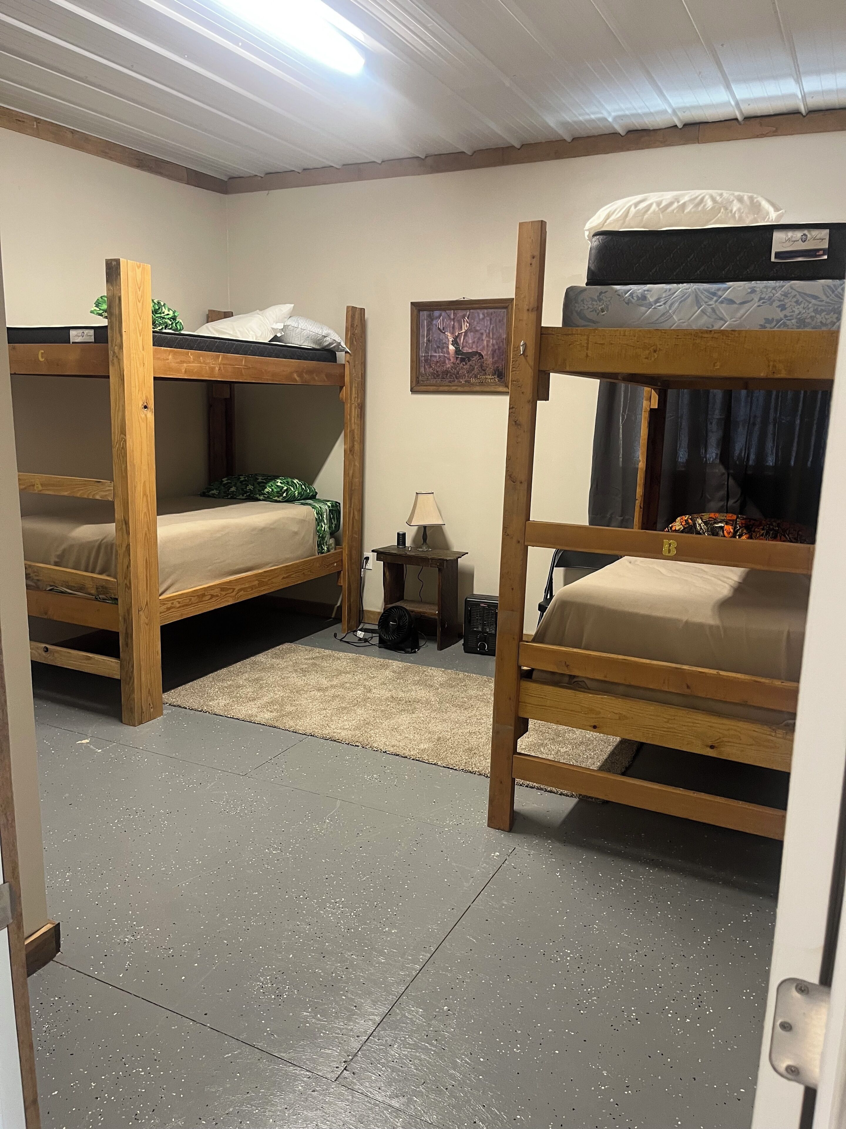 A bunk bed room with a framed deer picture on the wall, a small table with a lamp, and a fan on the floor.
