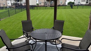 A patio with a round table and four chairs, surrounded by a black metal fence, with a grassy yard and residential buildings in the background. - Spacious Family Home• Sleeps 8 • Near Ft Campbell (Clarksville)