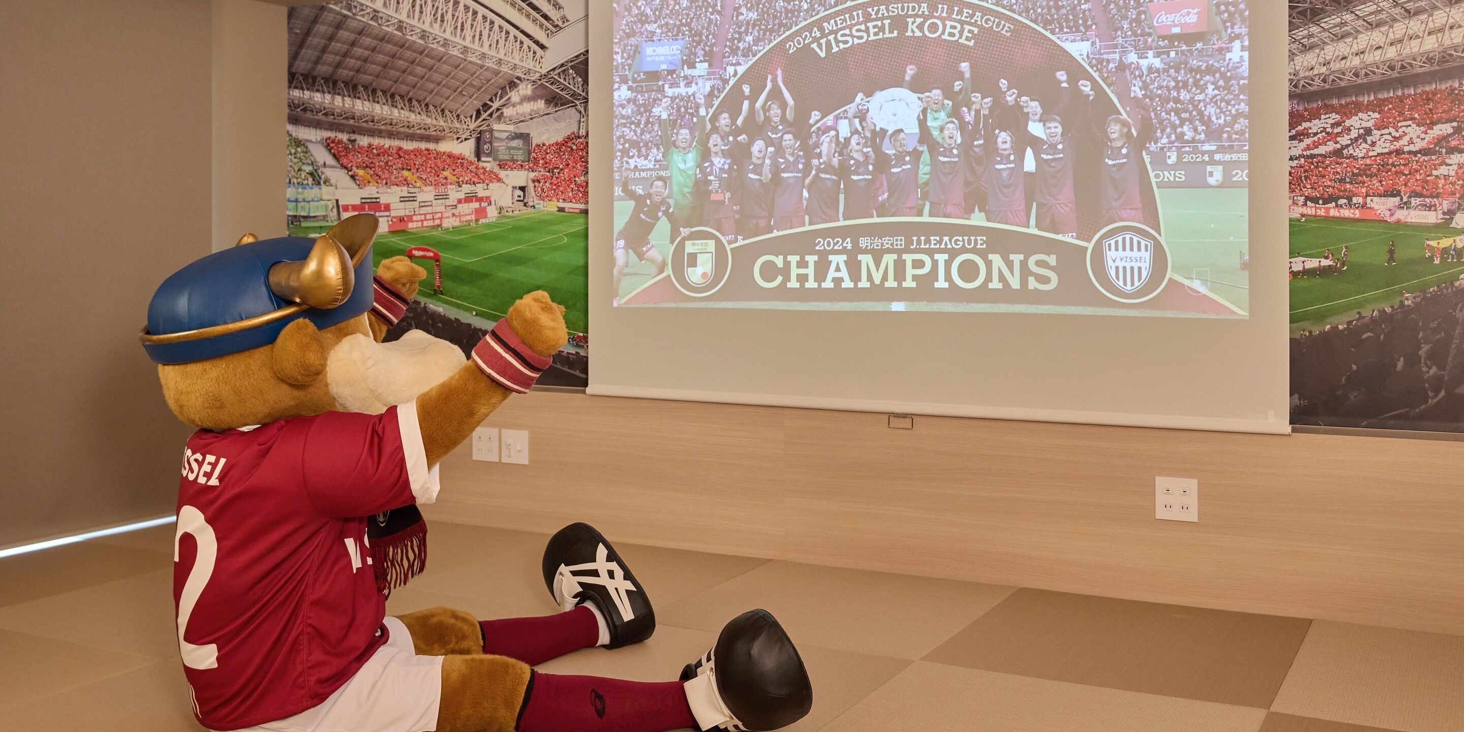 A mascot in a red jersey with the number 2, seated on the floor in a room with a large screen displaying a soccer match.