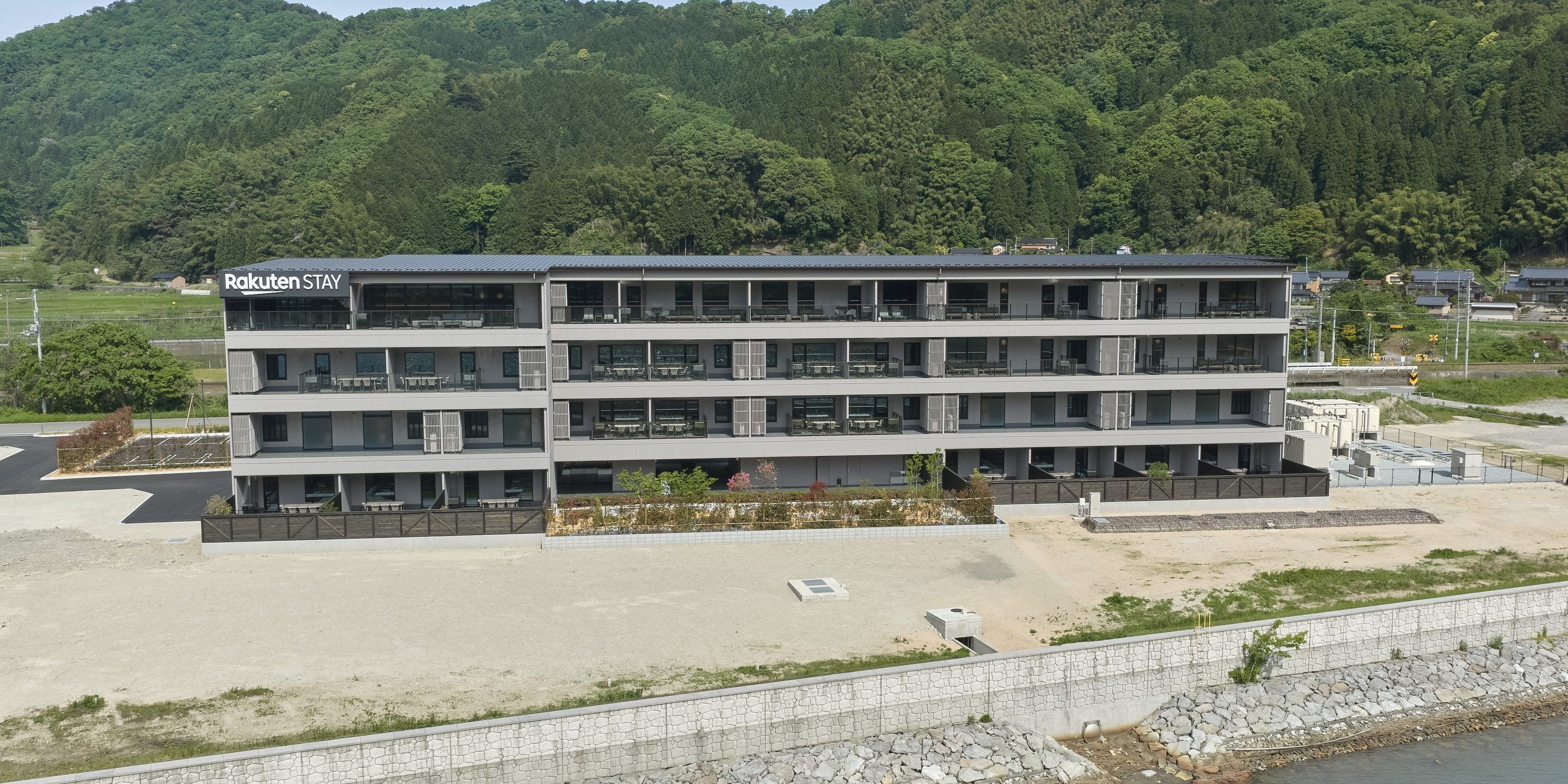 A multi-story building with a flat roof, labeled 'Rakuten STAY,' featuring balconies, a parking area, and a view of a forested hillside.