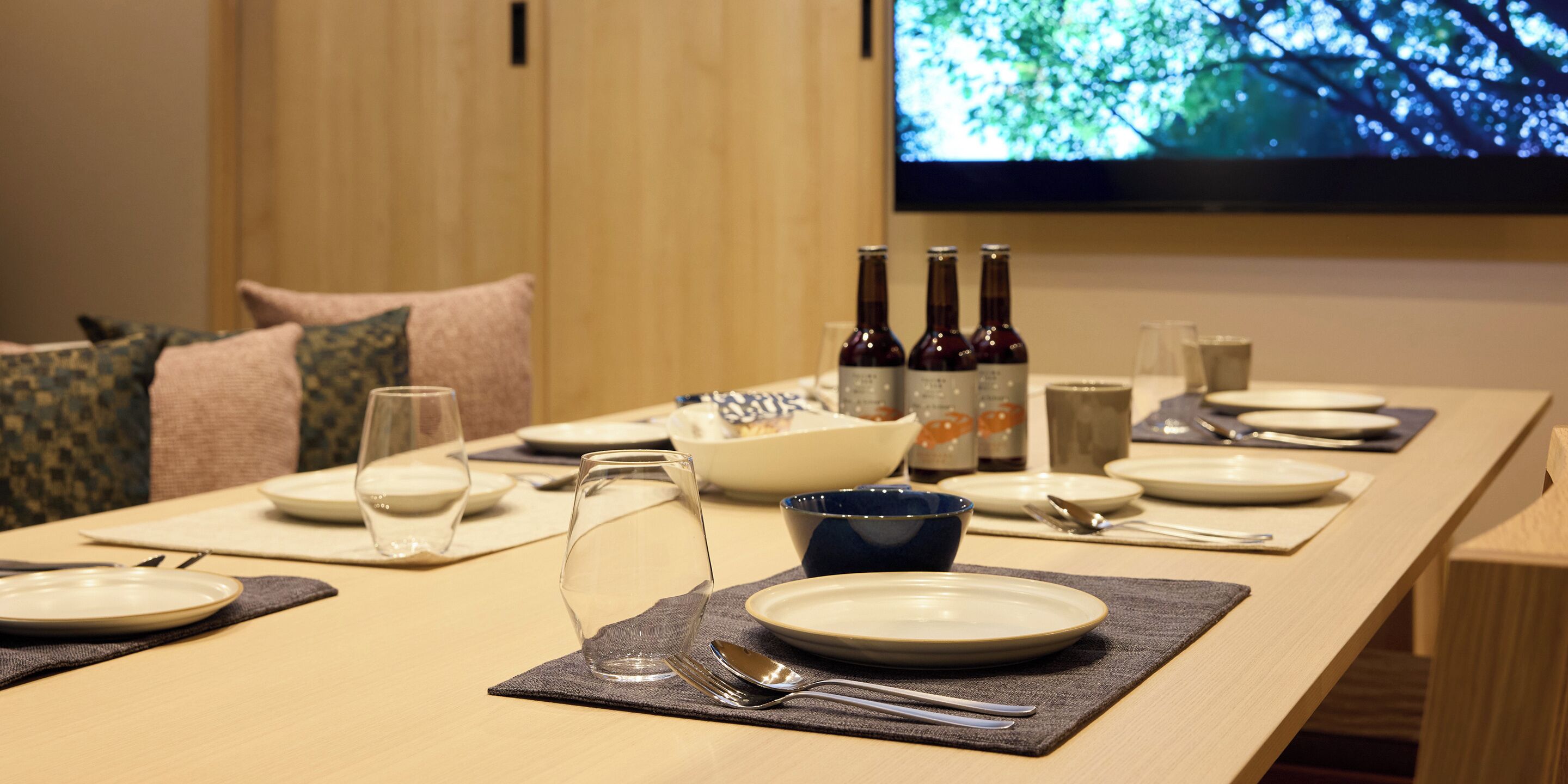 Een eettafel gedekt met borden, glazen en flessen, met een televisiescherm op de achtergrond.