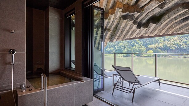 A modern bathroom with a glass-enclosed shower, a bathtub, and a balcony with a chair overlooking a lake.