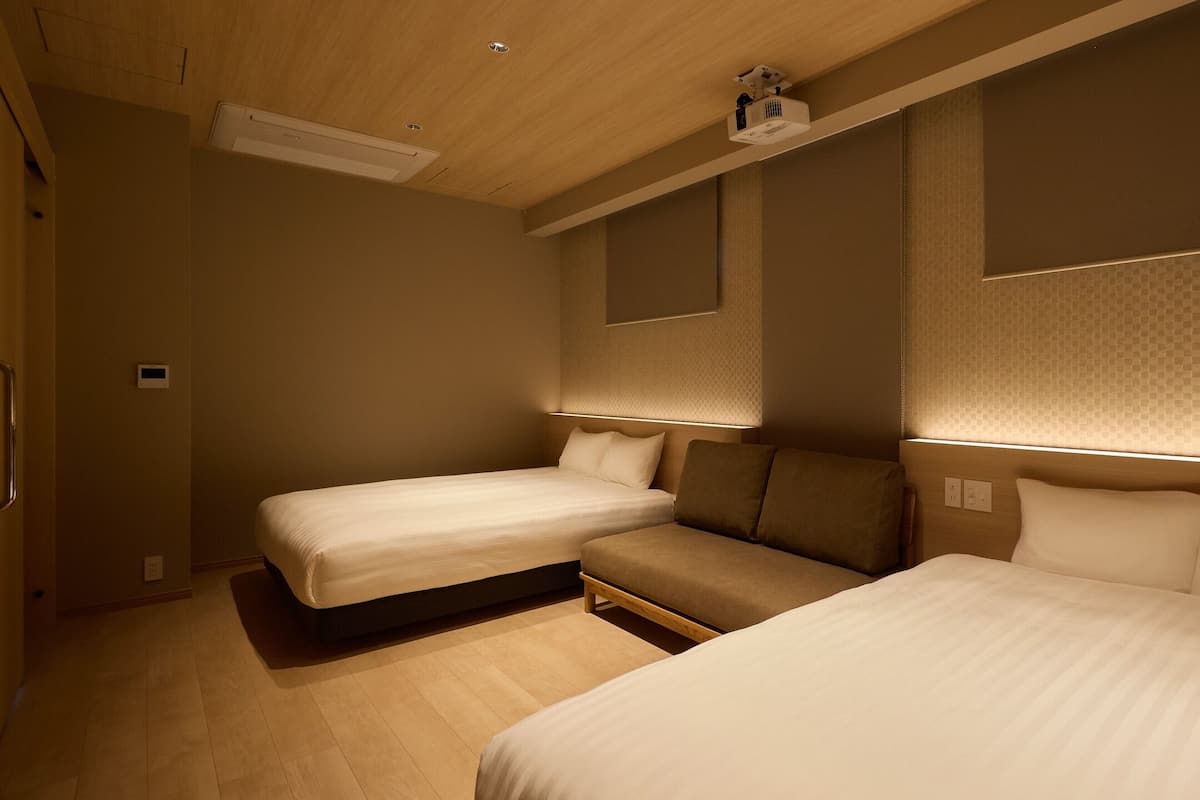 A hotel room with two beds, a sofa, a ceiling projector, and a wall-mounted light.
