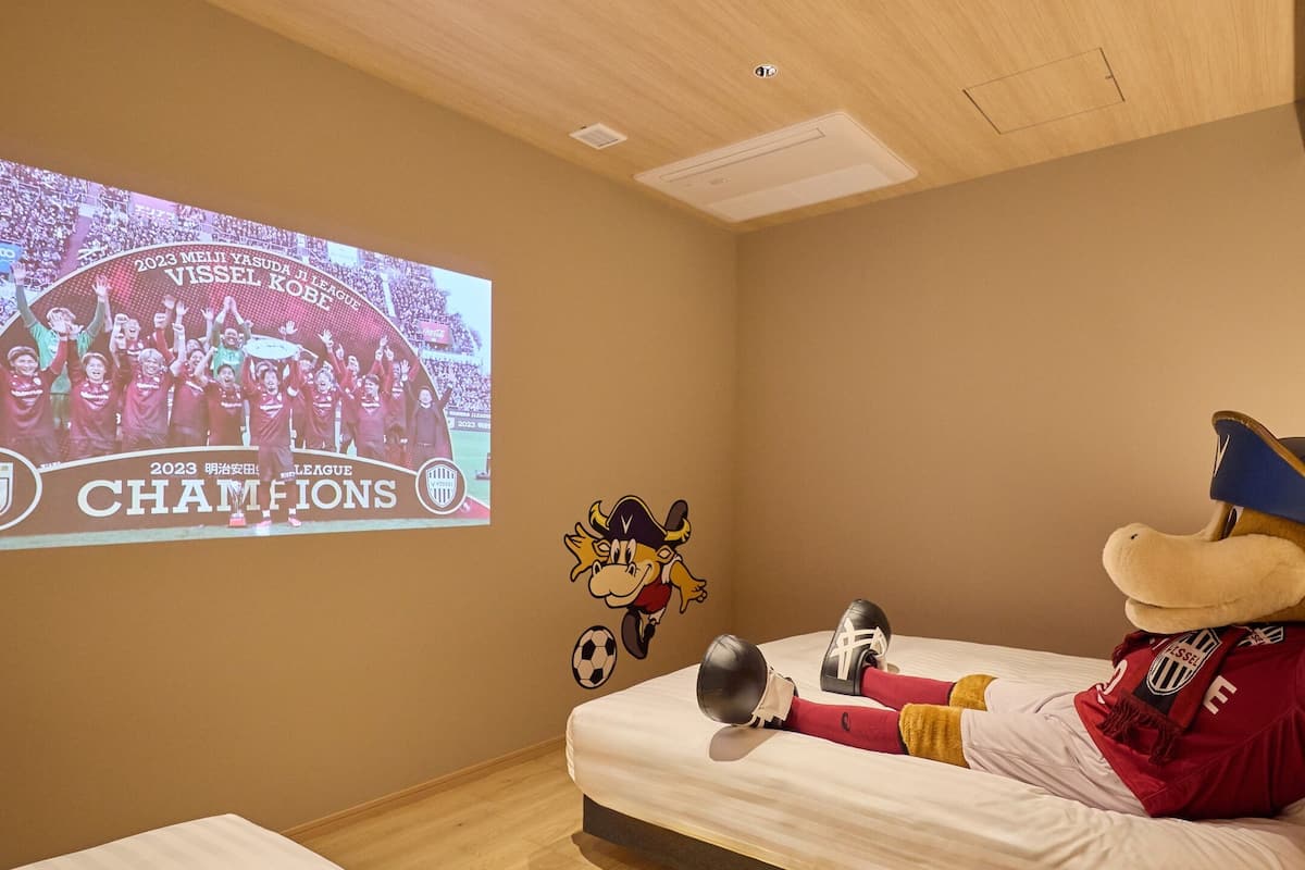A bedroom with a bed, a projection screen displaying a sports event, and a mascot figure.
