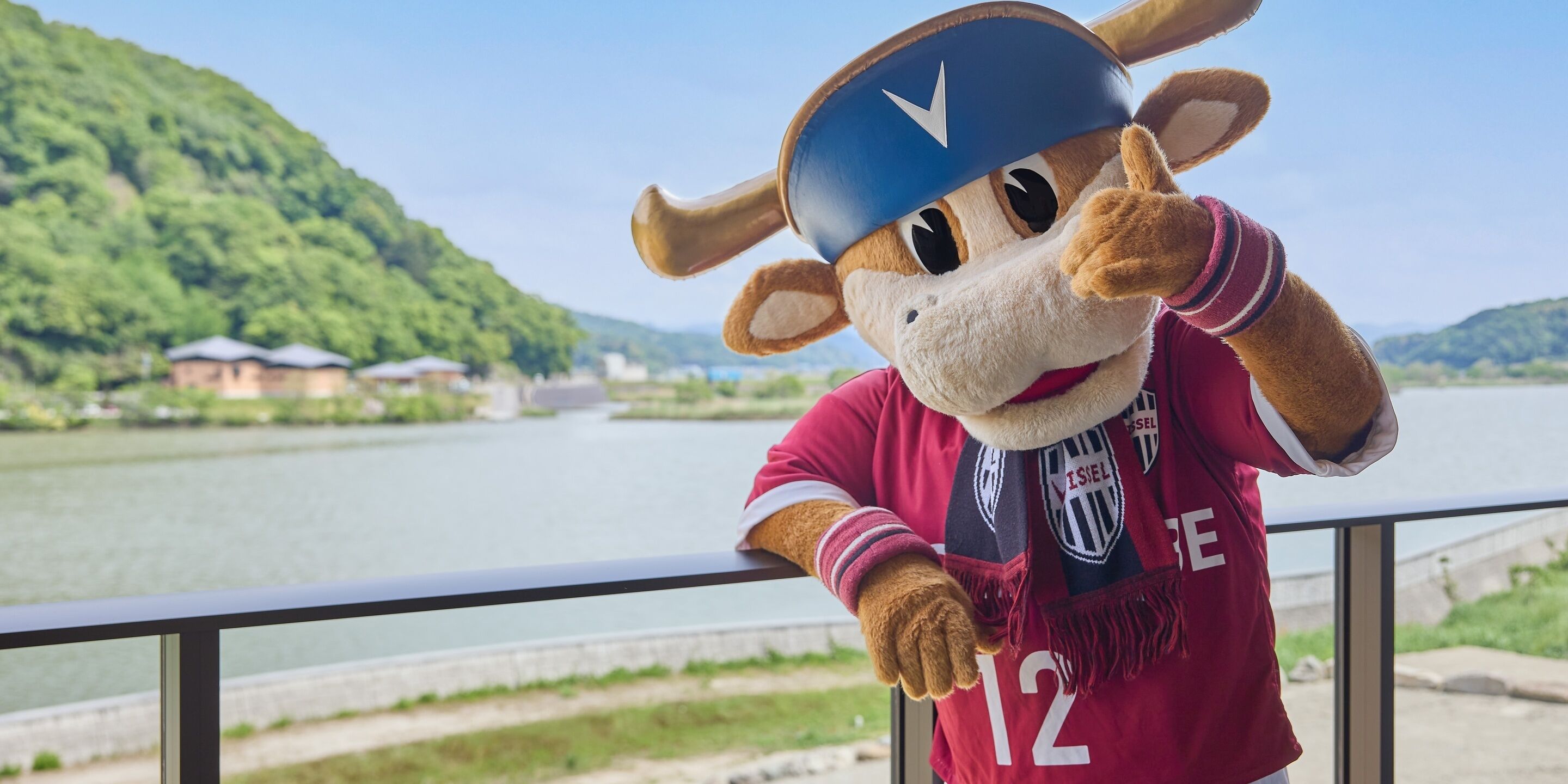A mascot wearing a red jersey with the number 12, standing by a railing with a river and greenery in the background.