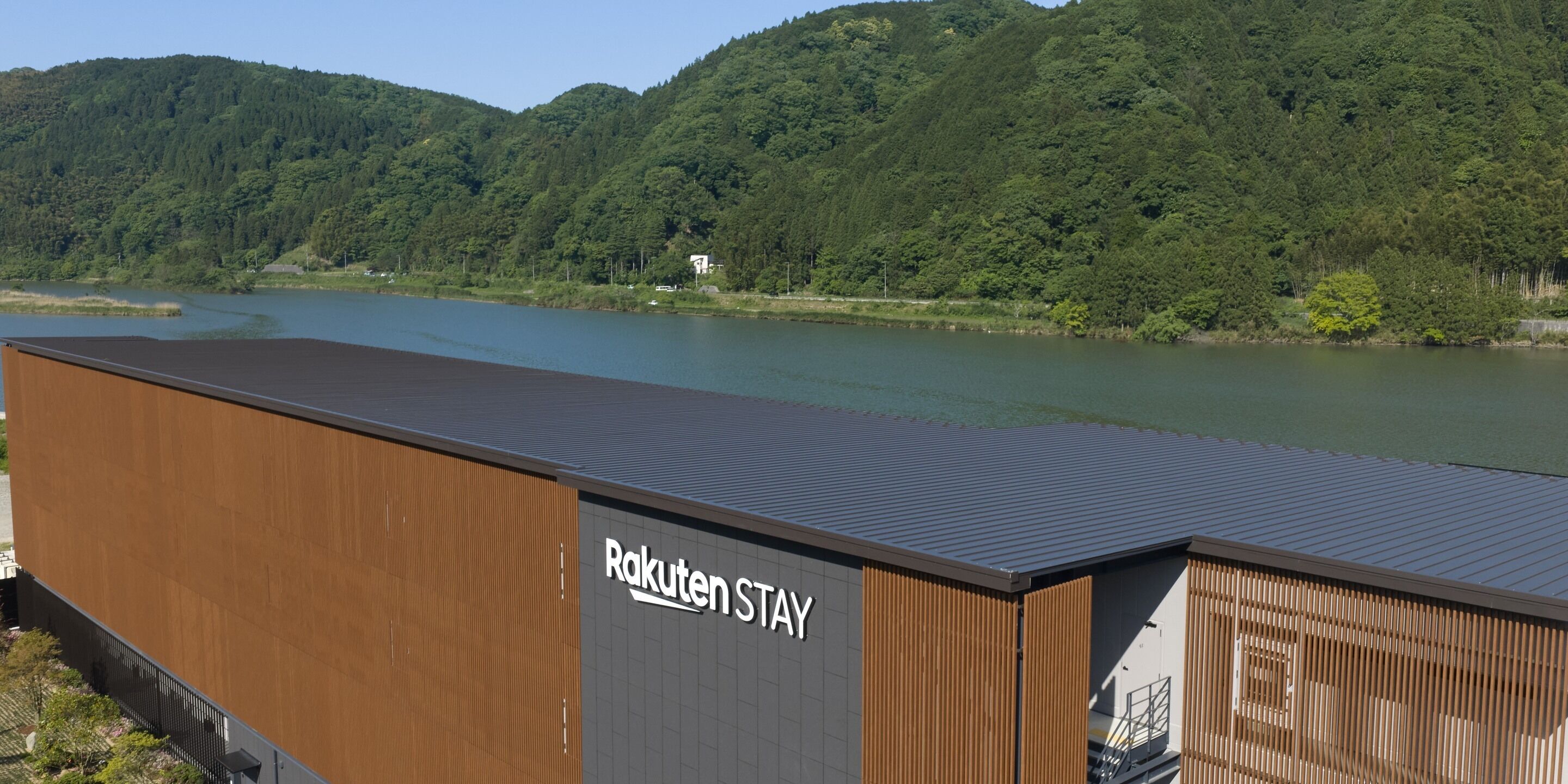Un edificio moderno con un cartel que dice 'Rakuten STAY' cerca de un lago con colinas boscosas al fondo.