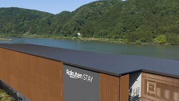 Un edificio moderno con un cartel que dice 'Rakuten STAY' cerca de un lago con colinas boscosas al fondo.