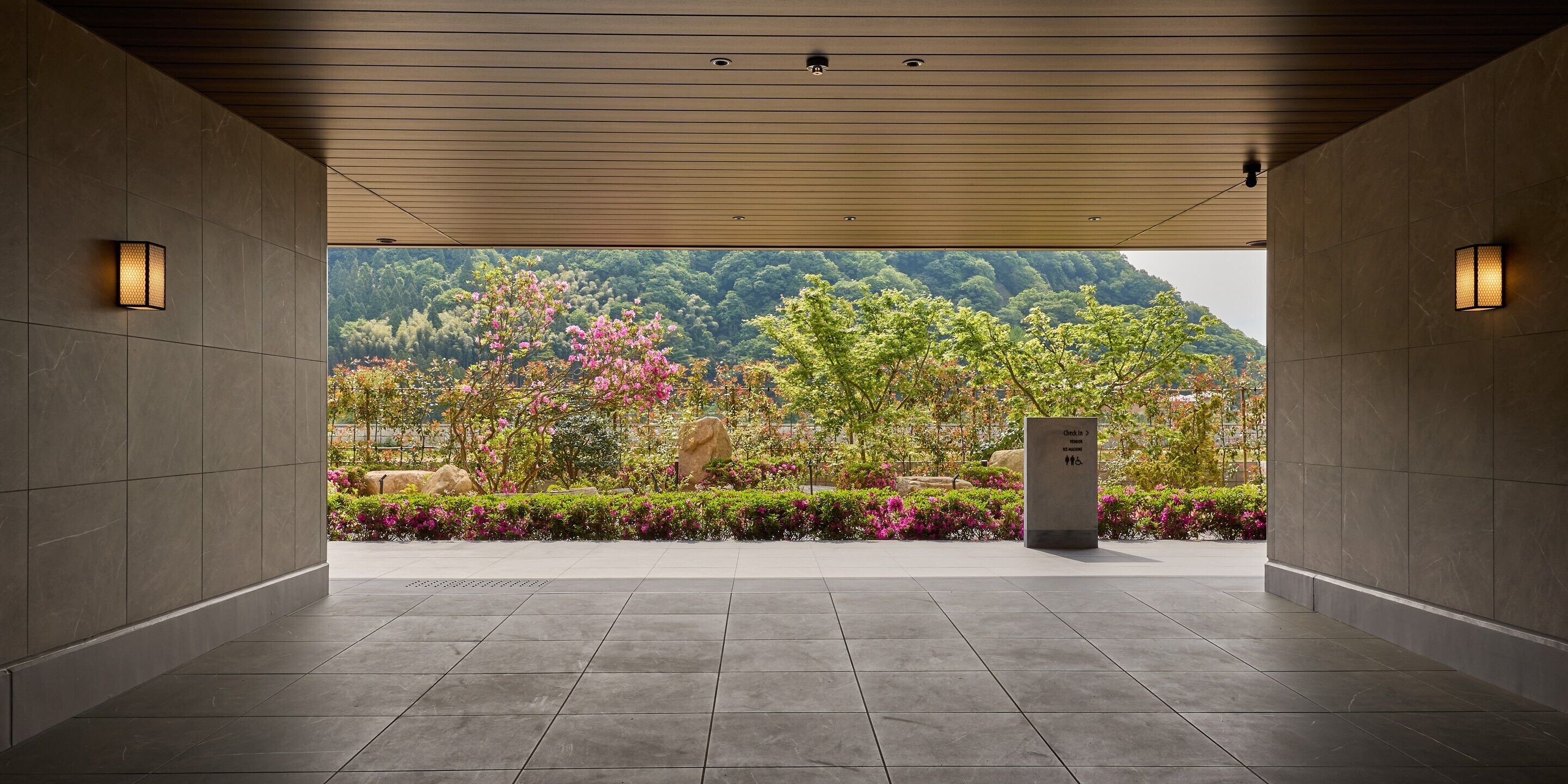 A covered walkway with tiled flooring, two wall-mounted lights, a stone pillar, and a view of a landscaped area with trees and flowers.