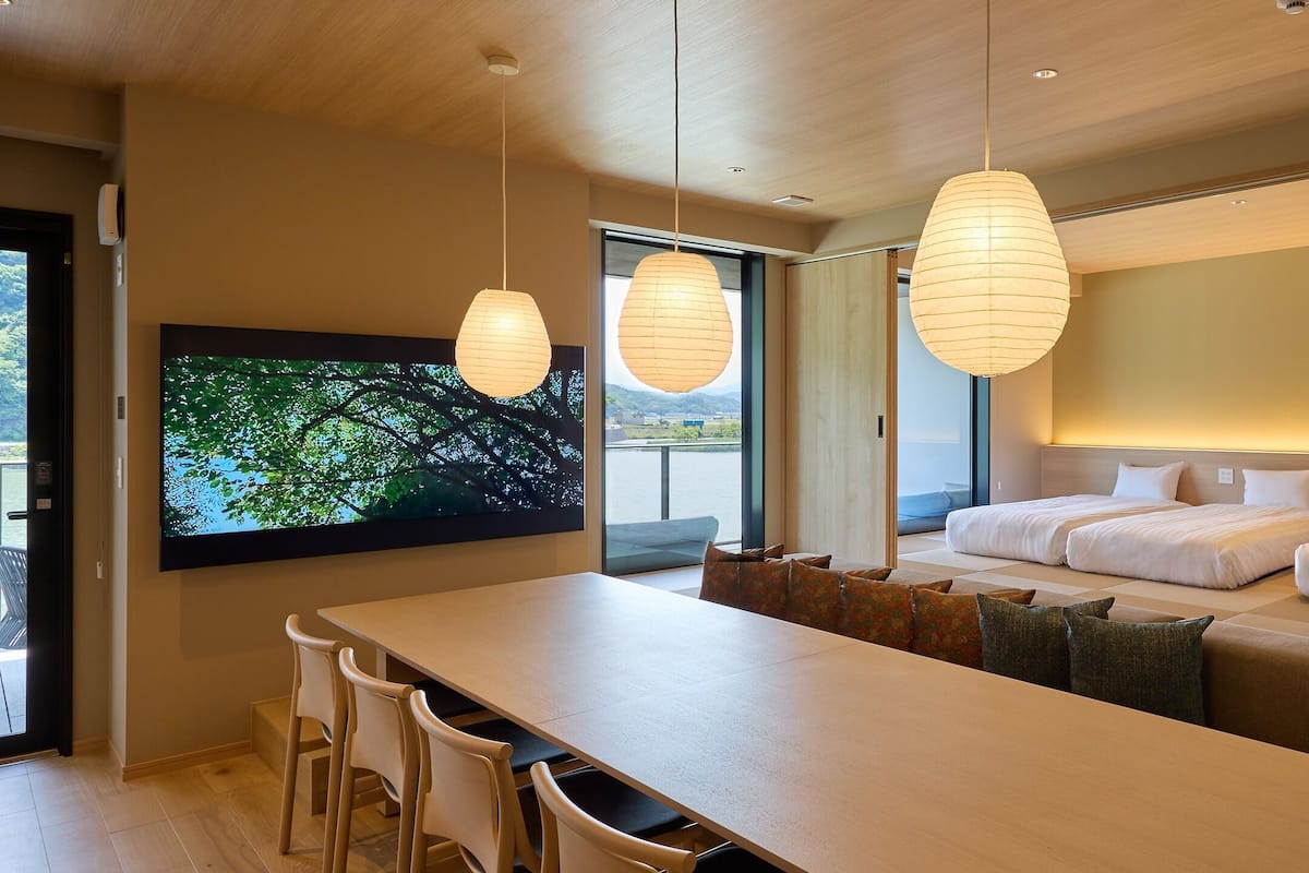 A modern hotel room with a large bed, a dining area with a long table and chairs, and a large TV screen displaying a nature scene.