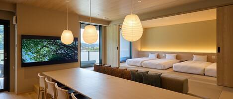 A modern hotel room with a large bed, a dining area with a long table and chairs, and a large TV screen displaying a nature scene.