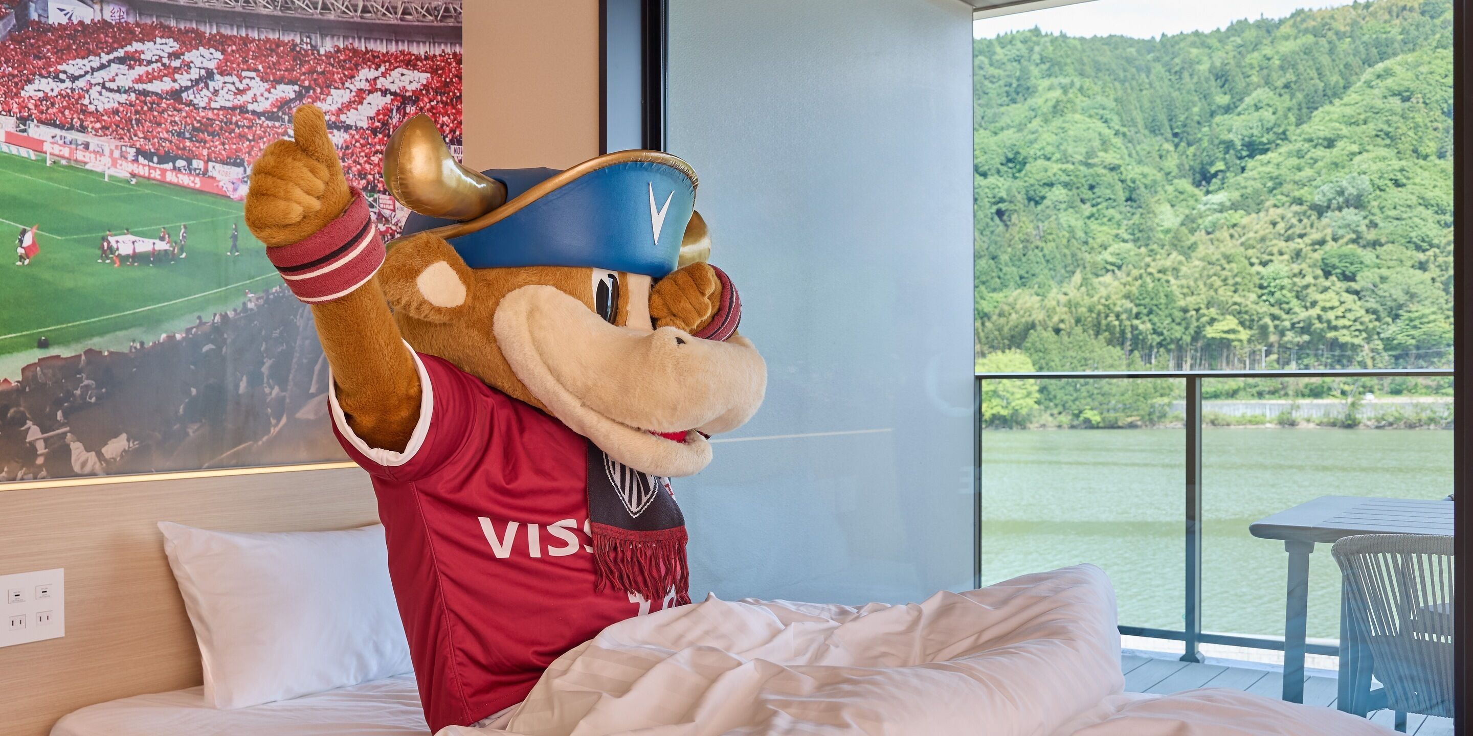 A mascot in a red jersey with a logo, a bed with white bedding, a window with a view of a forest and water, and a poster of a sports event.
