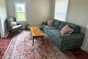 A living room with a green sofa, a wooden coffee table, a patterned rug, a chair, and a window. - The Traveler's Cottage - 15 minutes to sports park, downtown and Bourbon Trail (Elizabethtown)