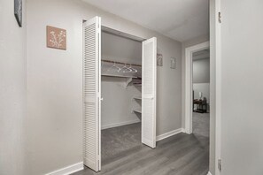 Ein begehbarer Kleiderschrank mit weißen, verleimten Türen, ein Holzboden und im angrenzenden Zimmer ein kleiner Tisch mit einer Lampe.