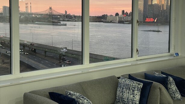 Een kamer met een groot raam dat uitkijkt op een stadsgezicht tijdens de zonsondergang, een bank met opvallende kussens, en een zichtbare weg met voertuigen.