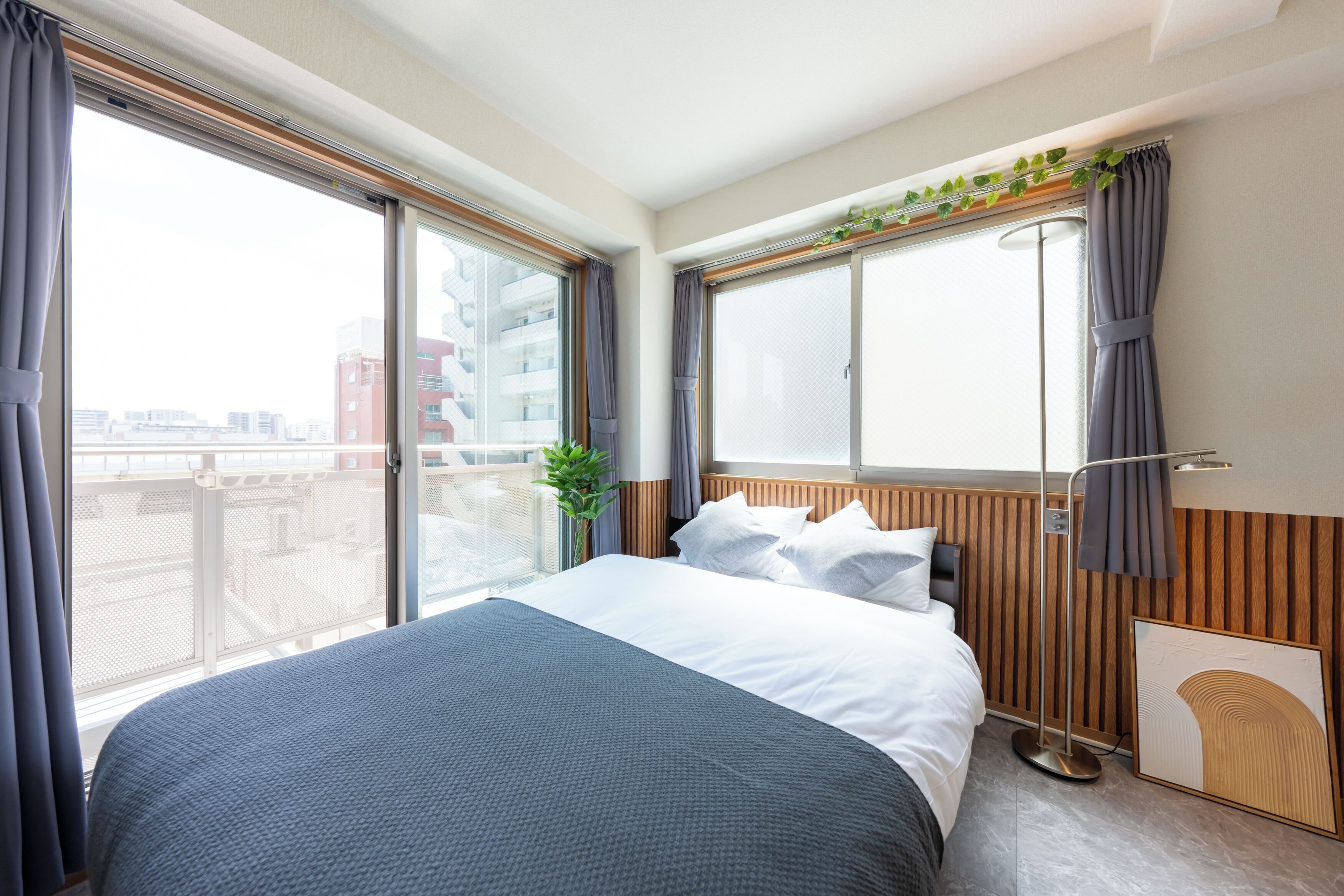 A hotel room with a large bed, a window with curtains, a plant, and a view of buildings outside.