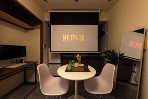 Uma sala de conferências com uma tela grande exibindo 'NETFLIX', uma mesa branca com duas cadeiras, uma pequena planta e um vaso com flores amarelas.