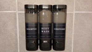 Tres botellas de productos de baño etiquetadas como champú, gel de ducha y acondicionador, sobre una pared de azulejos.