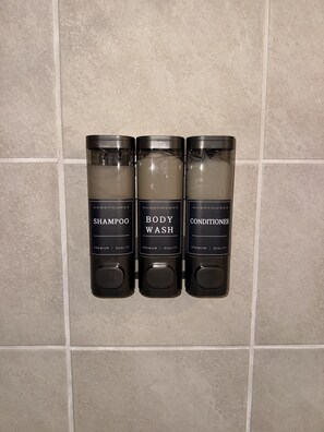 Tres botellas de productos de baño etiquetadas como champú, gel de ducha y acondicionador, sobre una pared de azulejos.