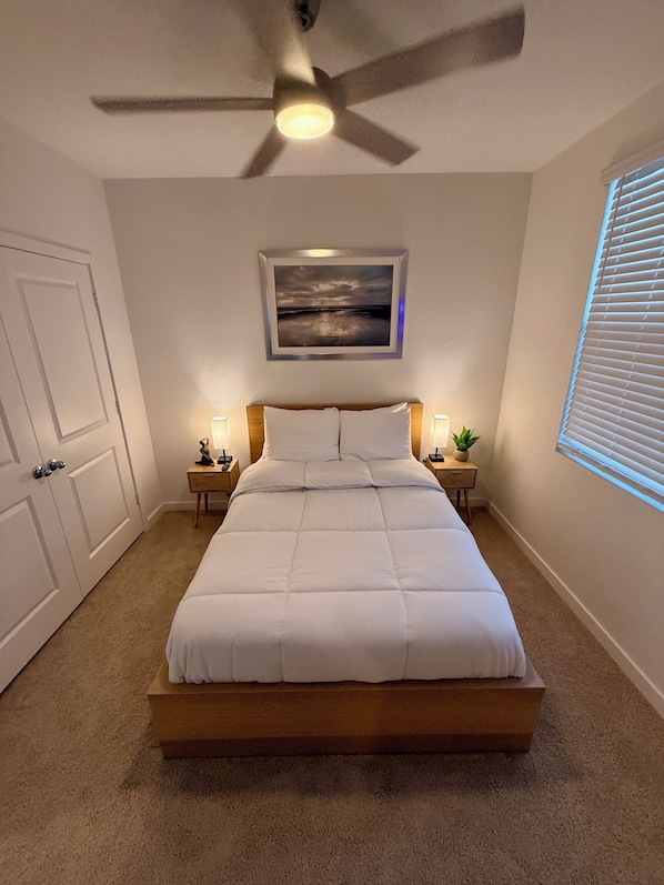 A bedroom with a bed, a ceiling fan, a framed picture on the wall, bedside tables with lamps, and a window with blinds.