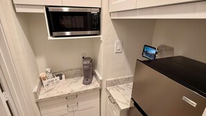 Cocina compacta con muebles blancos, microondas integrado, cafetera y refrigerador de acero inoxidable.