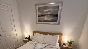 Una cama bien hecha con ropa blanca, un cuadro enmarcado encima y dos mesitas de noche con lámparas y una planta en maceta.