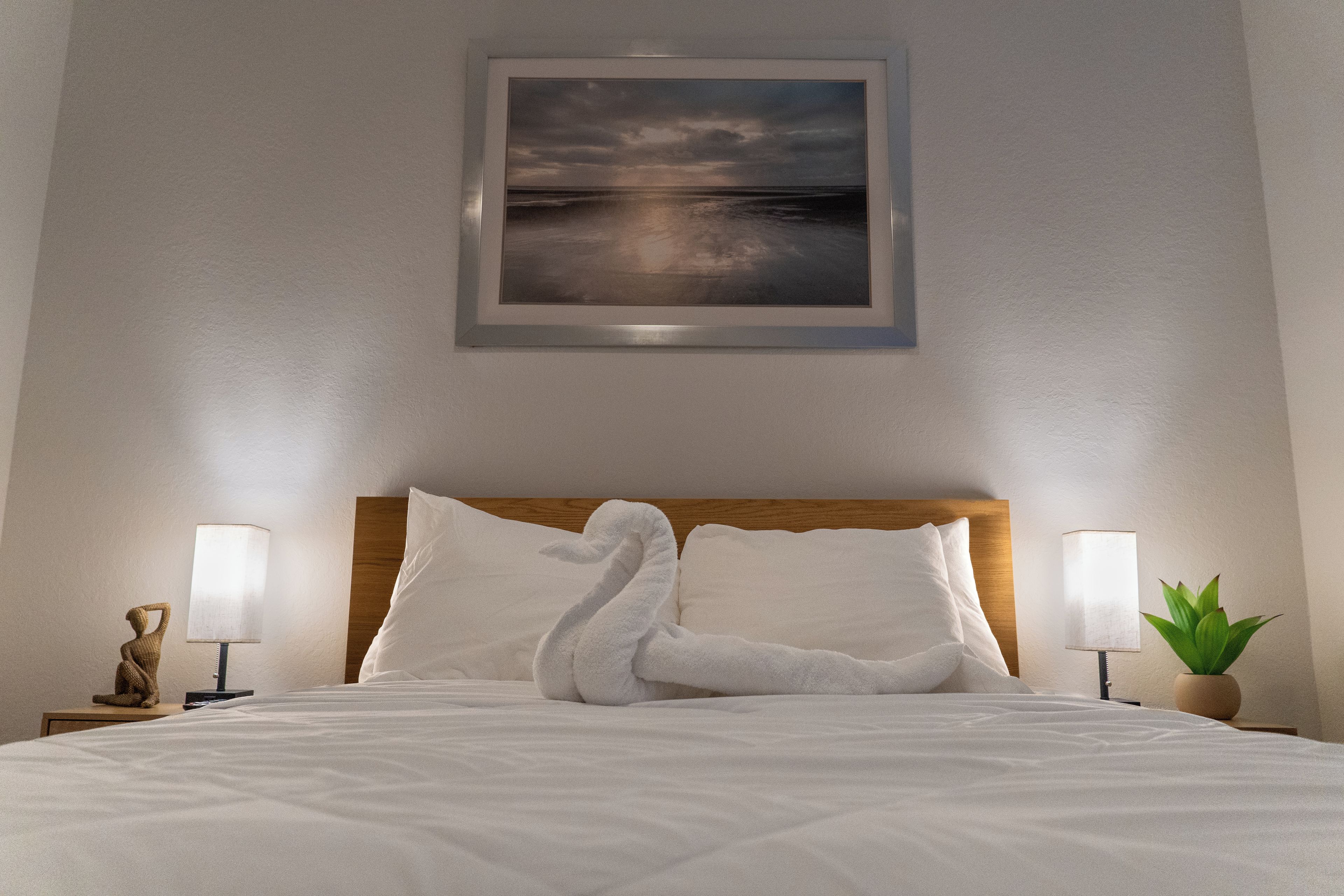 Ein ordentlich bezogenes Bett mit weißer Daunendecke, zwei Kissen und einer dekorativen Schwan-Handtuch-Arrangement. An der Wand über dem Bett hängt ein gerahmtes Landschaftsbild.