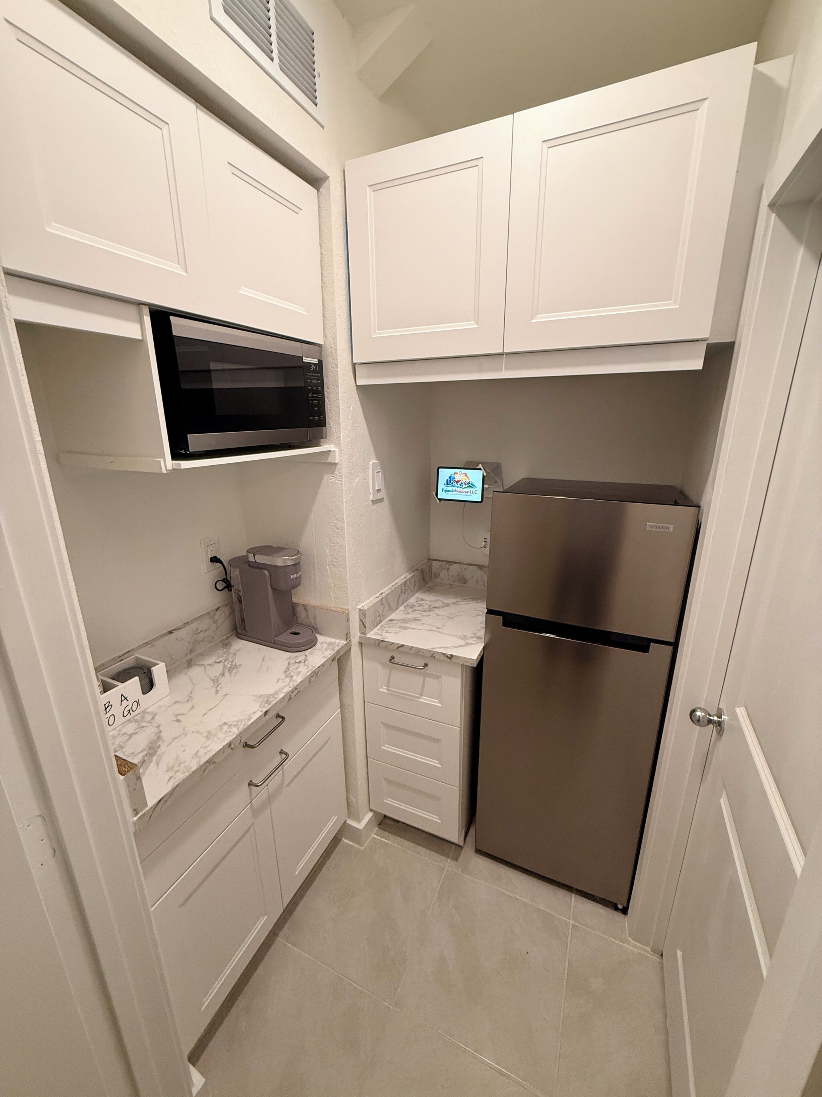 Una cocina compacta con armarios blancos, un refrigerador de acero inoxidable, un microondas y una cafetera.