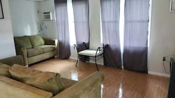 En stue med sofa, stol og lille bord. Gardiner er der ved vinduerne.