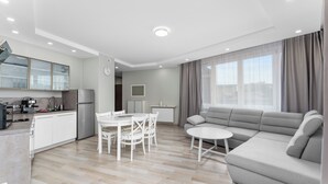 Een moderne woonkamer met een bank, een eettafel met stoelen, een keuken met kasten en apparatuur, en een groot raam met gordijnen.