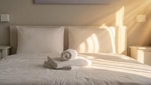 Een net opgemaakt bed met opgerolde handdoeken, een eenvoudig hoofdbord en een wandlamp.