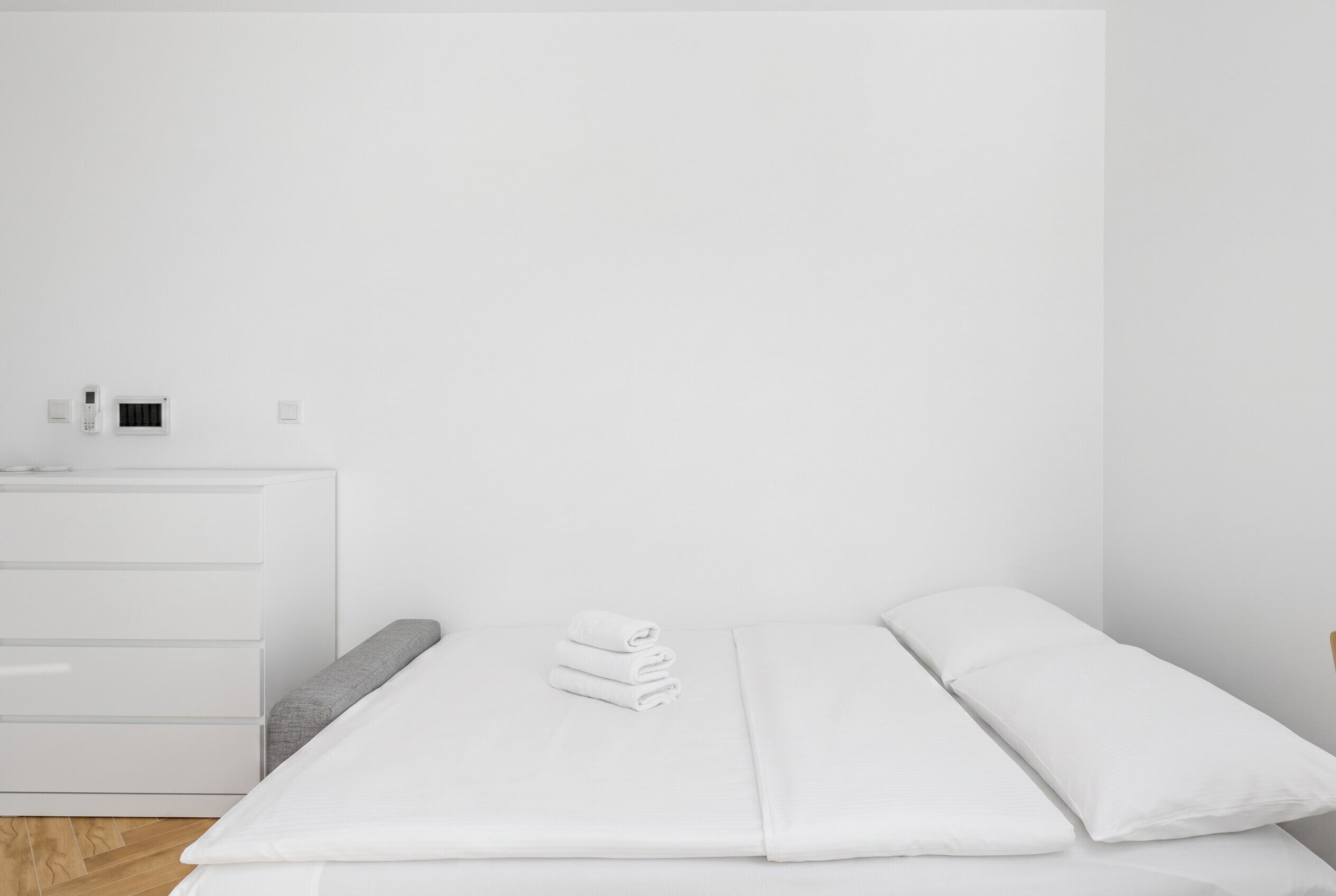 Une chambre d’hôtel minimaliste, avec un lit, une commode blanche et un climatiseur fixé au mur.