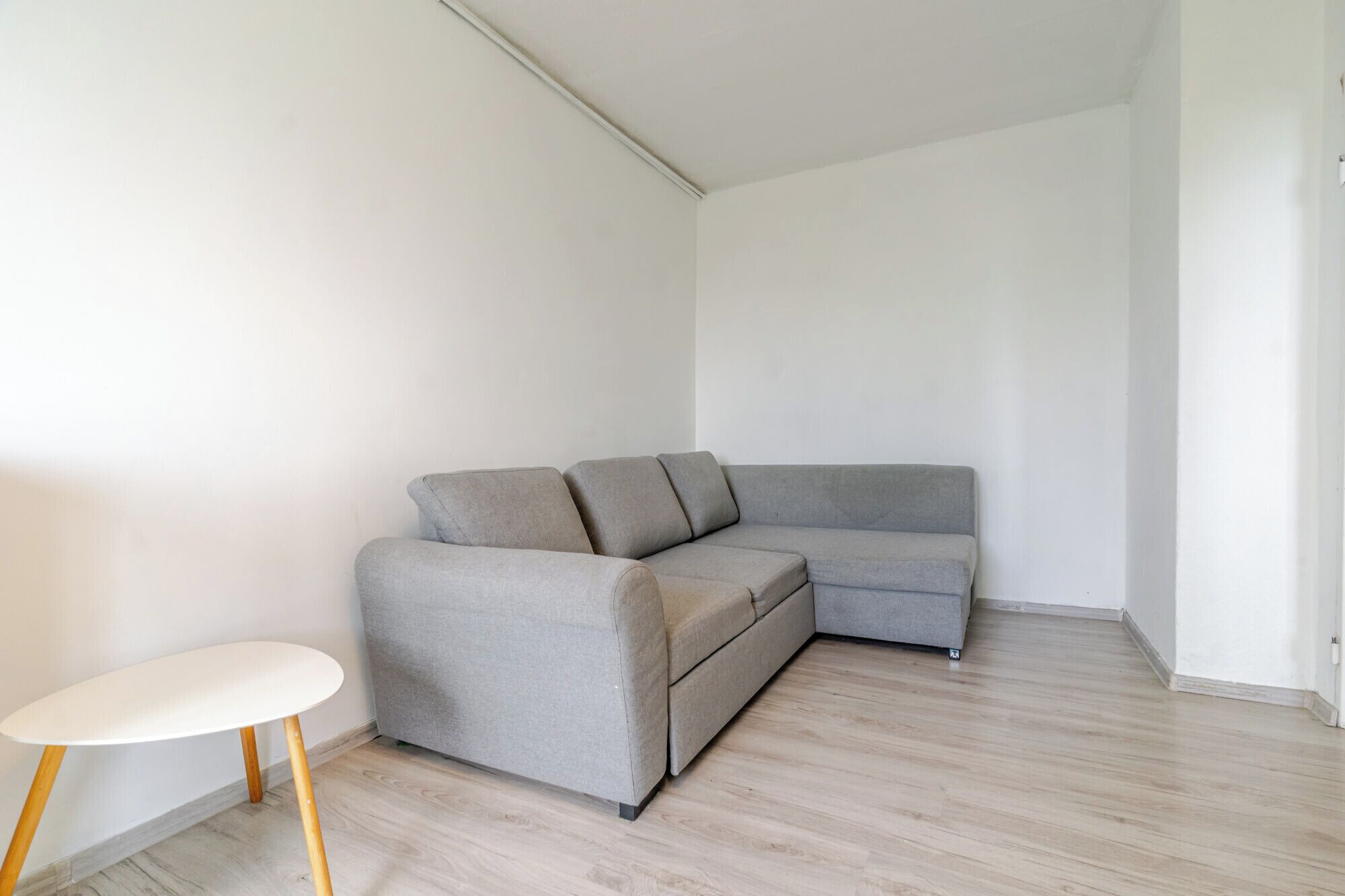 Quarto de canto com um sofá cinzento, uma mesa pequena branca e soalho de madeira.