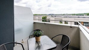 Un balcon avec une table en verre et deux chaises, un pot de fleurs et une vue sur des bâtiments et de la verdure.