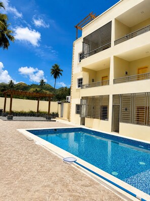 A multi-story building with a swimming pool, balconies, and palm trees in the background.