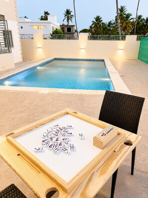 Un espace aménagé au bord de la piscine, avec une table préparée pour jouer aux dominos, une chaise et une vue sur un bâtiment et des palmiers.