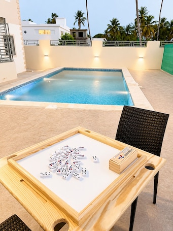 A poolside area with a table set for dominoes, a chair, and a view of a building and palm trees.. Samaná Paradise • Pool, Ocean Views & Beachside Living in Los Robalos