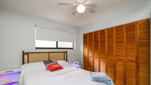Ein Schlafzimmer mit Bett, Deckenventilator, Fenster mit Jalousien, einem Holzschrank und einem Nachttisch mit Lampe.