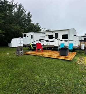 A Sprinter Select RV with a wooden deck, two blue chairs, a red chair, and a black table. - Newly Renovated Trailer, Sleep 8 Minutes to Beach & Amenities (Beaubassin East)