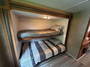 A bunk bed with a headboard, a lower bed with a striped blanket, and a ceiling light fixture. - Newly Renovated Trailer, Sleep 8 Minutes to Beach & Amenities (Beaubassin East)
