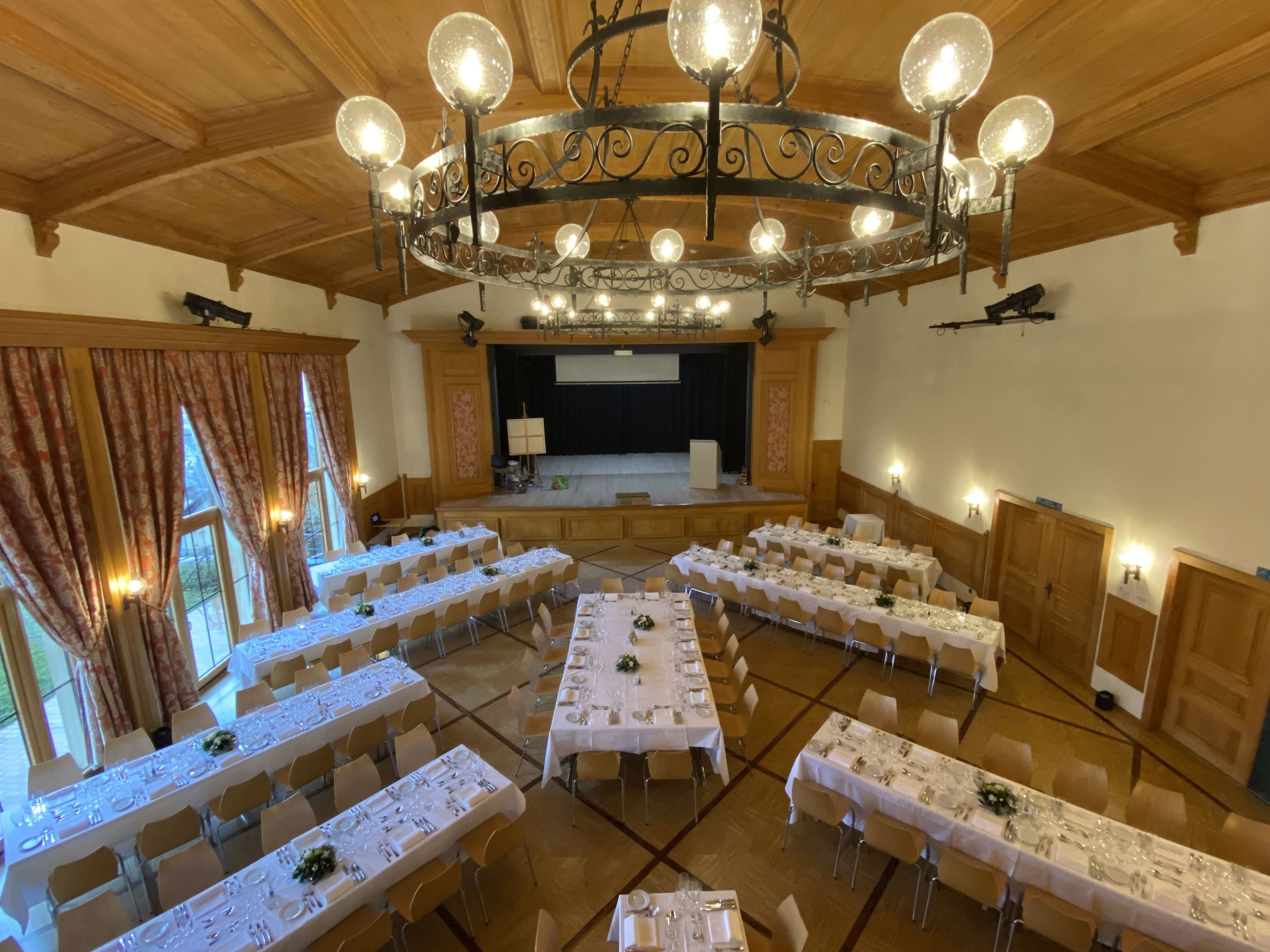 A banquet hall with multiple round tables set for dining, a stage with a screen, and a decorative chandelier.