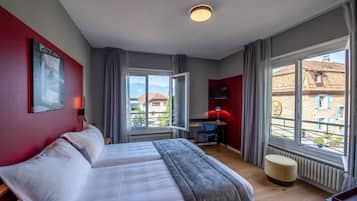 A hotel room with a bed, a desk, a chair, and a view of buildings through large windows.