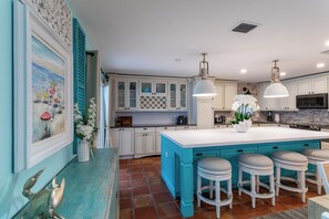 Cozinha com armários brancos, uma ilha grande com banquetas, iluminação pendente e um quadro na parede.